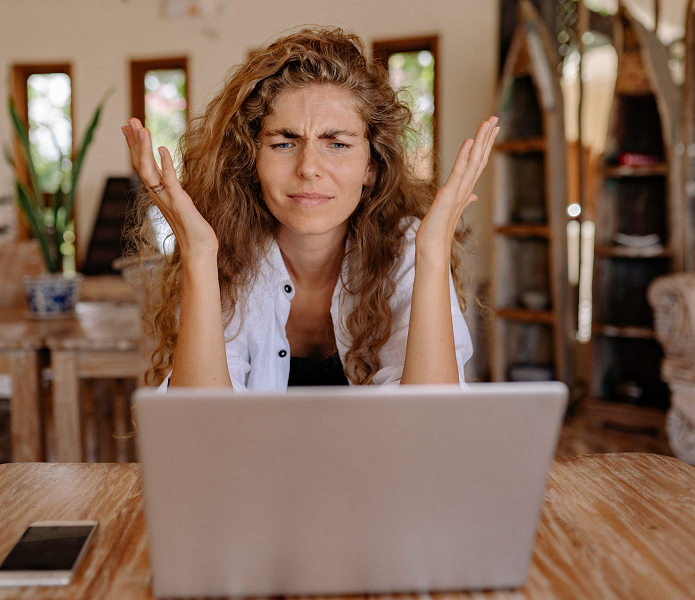 A woman sitting in font of her laptop with her hands in the air. She looks frustrated. 